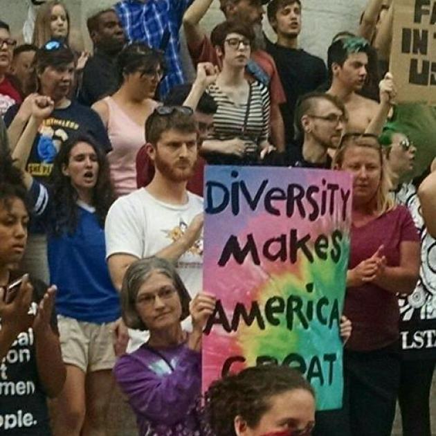 Person holding sign in crowd of people with fists in air, reading Diversity Makes America Great against tie-dyed background