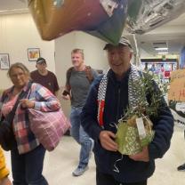 Man walking through airport holding a plant with people and balloons surrounding him